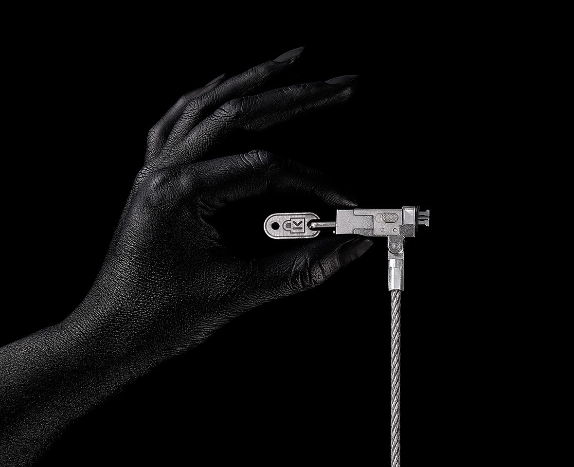 A human hand holding a Kensington Slim NanoSaver EQ Keyed Laptop Lock on a dark background.